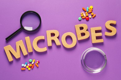Word Microbes made with wooden letters , magnifying glass and pills on purple background, flat lay