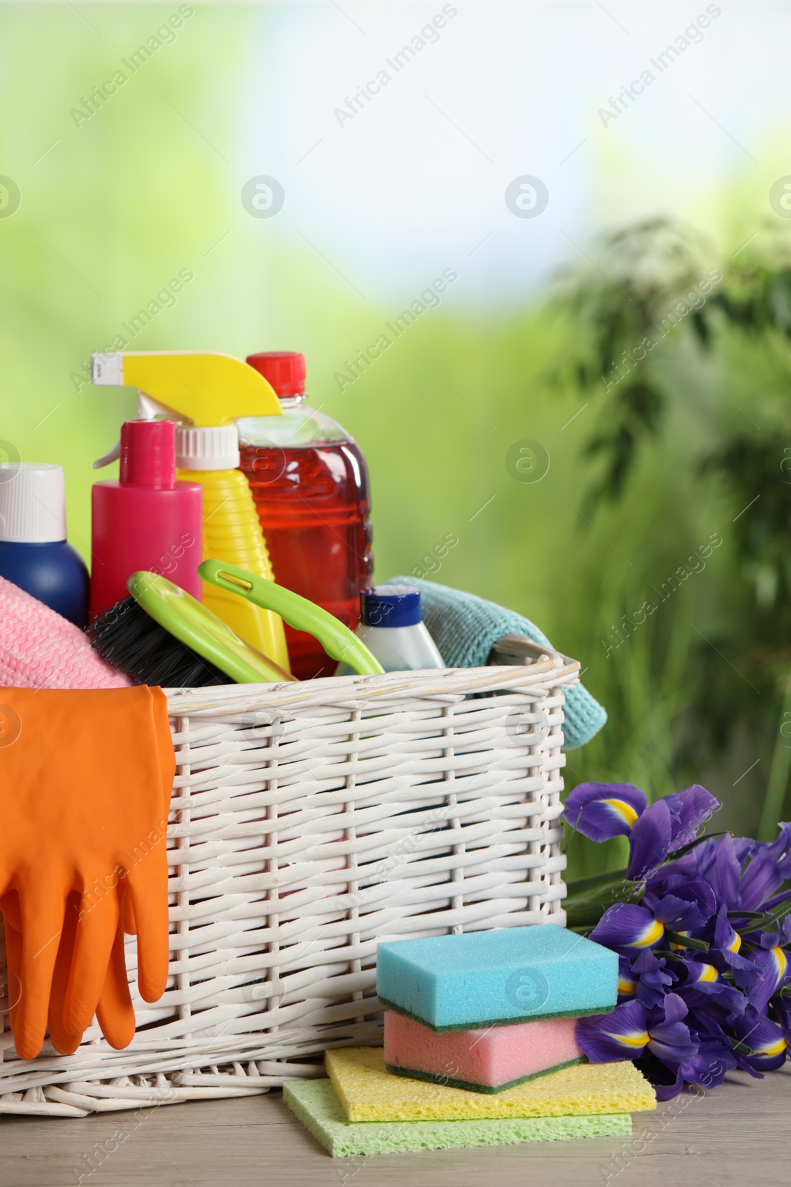 Photo of Spring cleaning. Plastic basket with detergents, supplies and beautiful flowers on wooden table outdoors