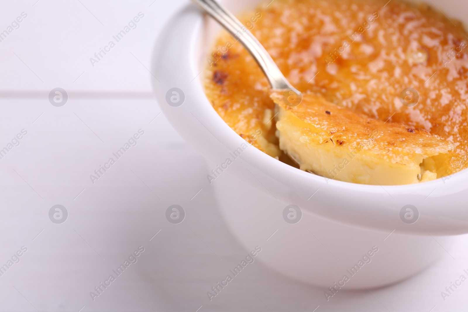 Photo of Taking delicious creme brulee with spoon from bowl at white wooden table, closeup. Space for text