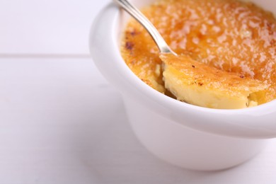 Taking delicious creme brulee with spoon from bowl at white wooden table, closeup. Space for text