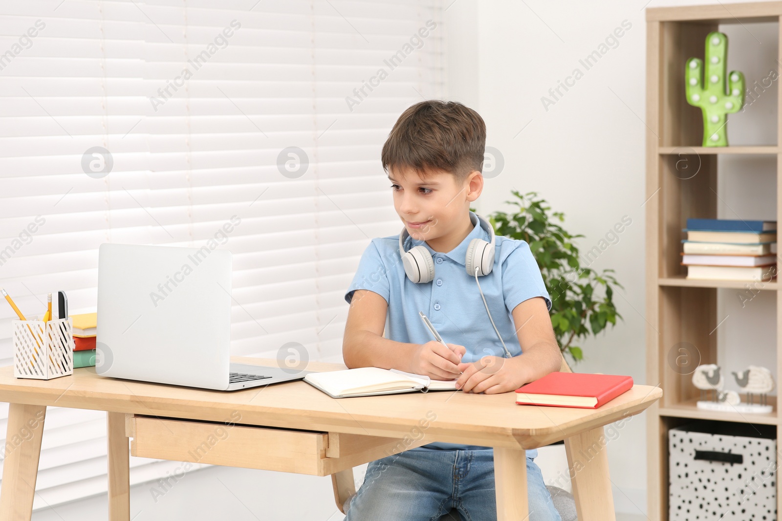 Photo of Boy writing in notepad near laptop at desk in room. Home workplace