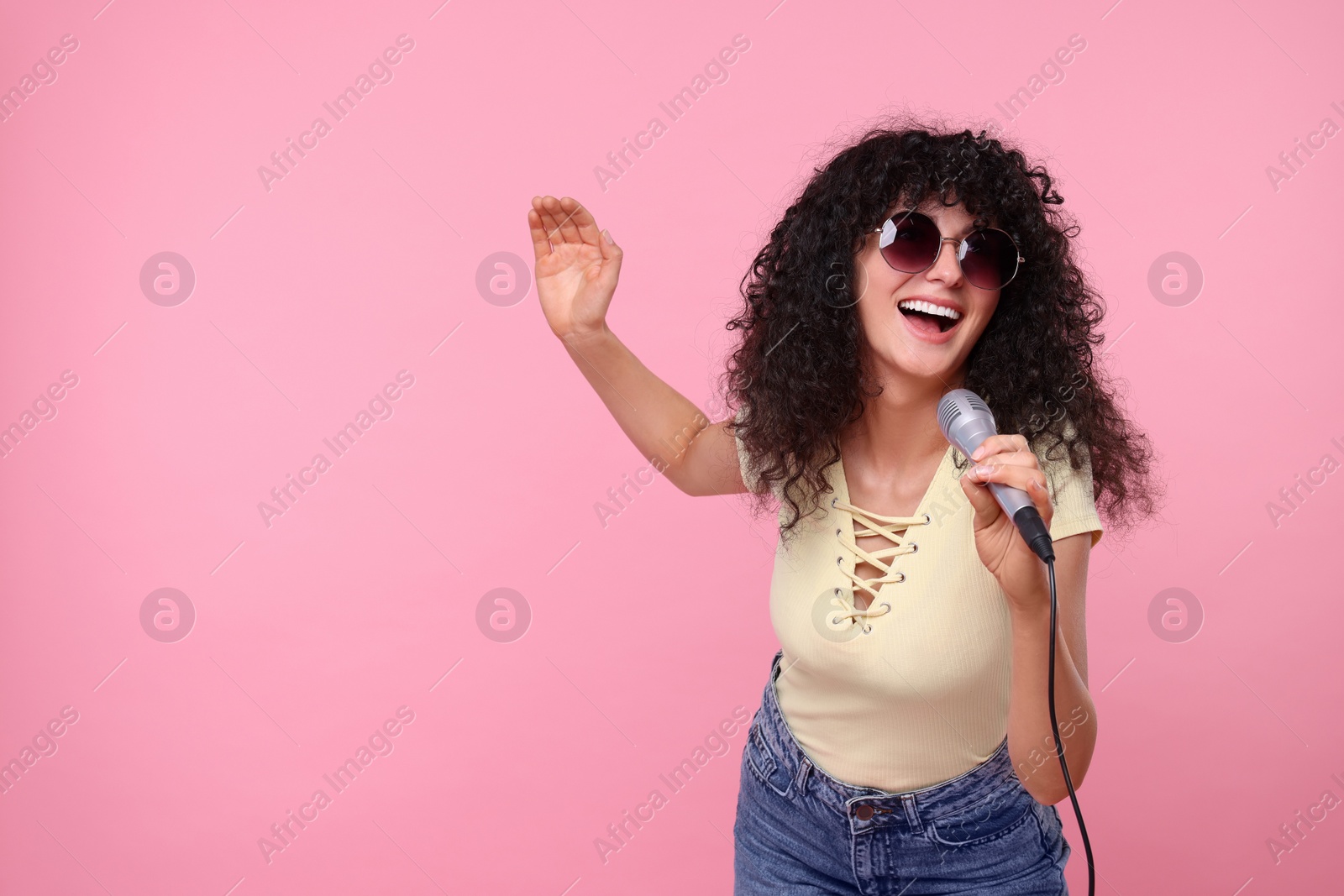 Photo of Beautiful young woman with microphone and sunglasses singing on pink background. Space for text