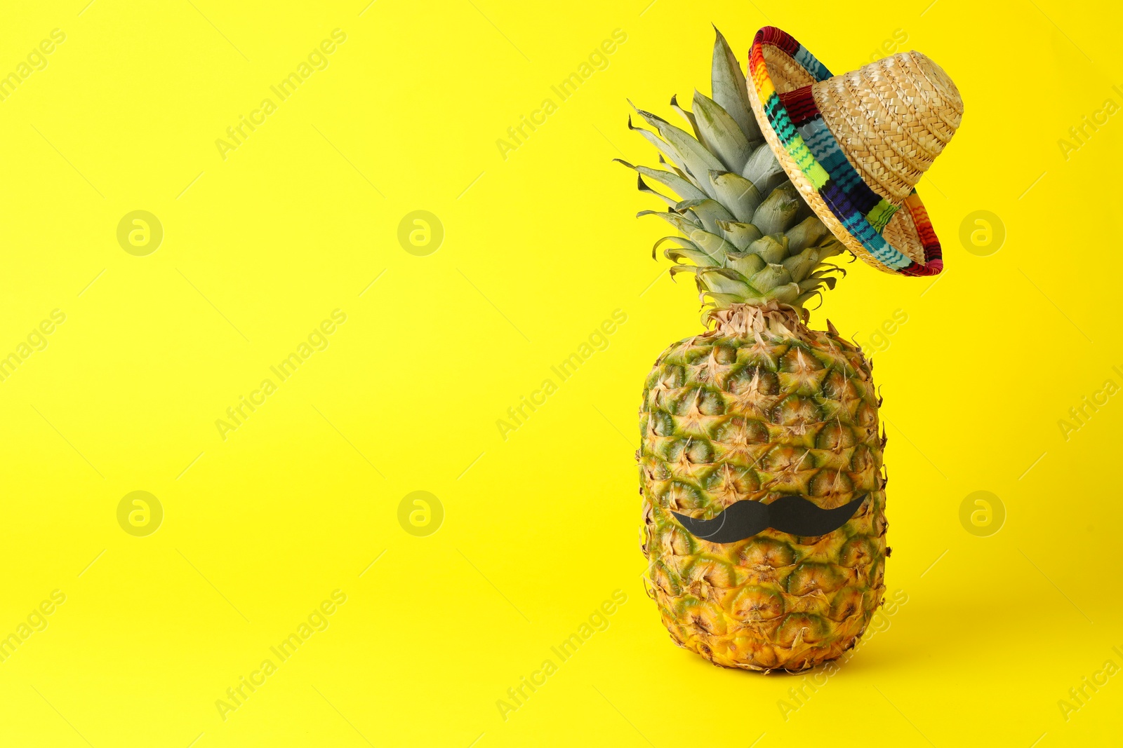 Photo of Pineapple with Mexican sombrero hat and fake mustache on yellow background, space for text