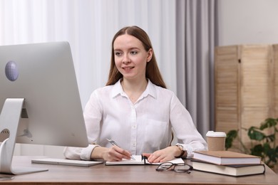 Photo of E-learning. Young woman taking notes during online lesson at wooden table indoors