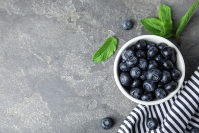 Photo of Bowl of tasty fresh blueberries, leaves and fabric on grey stone surface, flat lay. Space for text