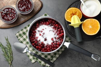 Photo of Making cranberry sauce. Fresh cranberries with sugar in saucepan and ingredients on gray table, flat lay