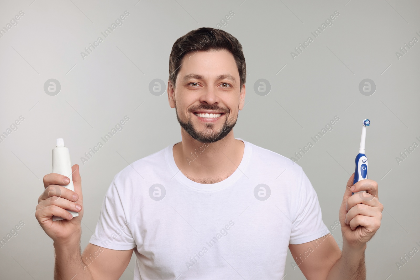 Photo of Happy man holding electric toothbrush and tube of toothpaste on light grey background