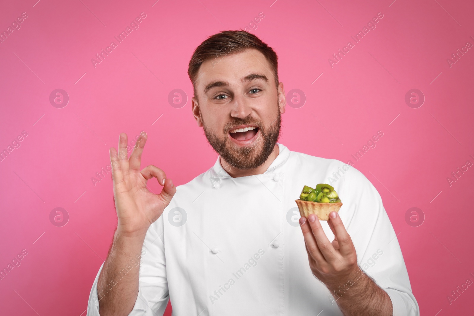 Photo of Happy professional confectioner in uniform with delicious tartlet showing okay gesture on pink background