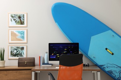 Stylish workplace with modern computer and SUP board near light wall in room. Interior design