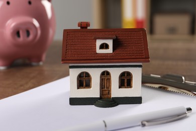 Photo of House model, clipboard, pen and piggy bank on wooden table indoors, selective focus