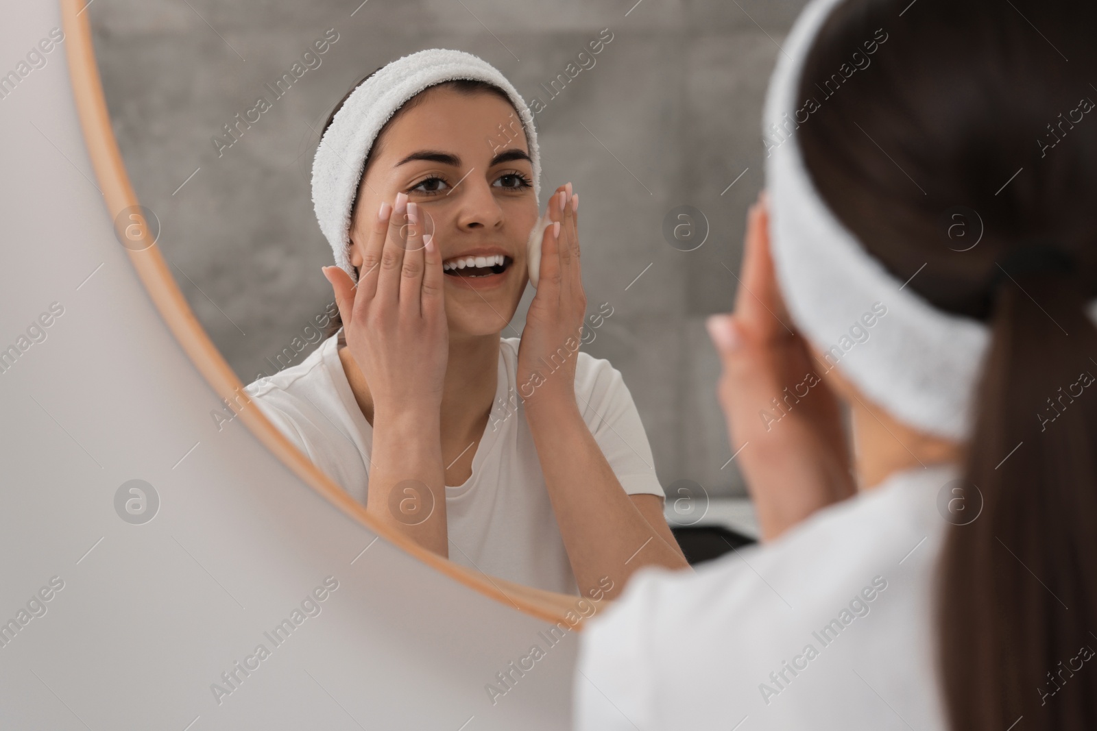 Photo of Young woman with headband washing her face near mirror in bathroom