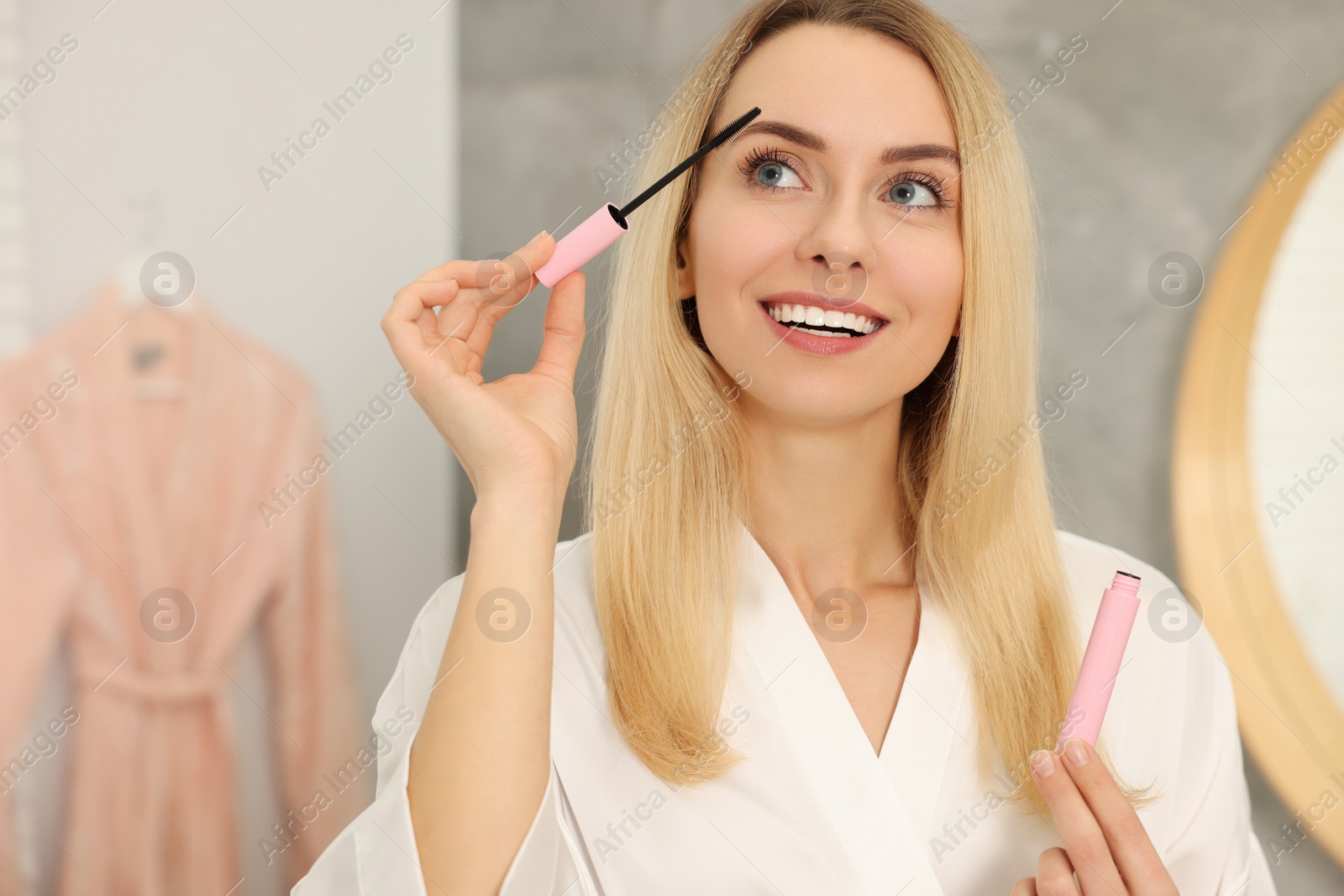 Photo of Beautiful happy woman applying mascara in bathroom