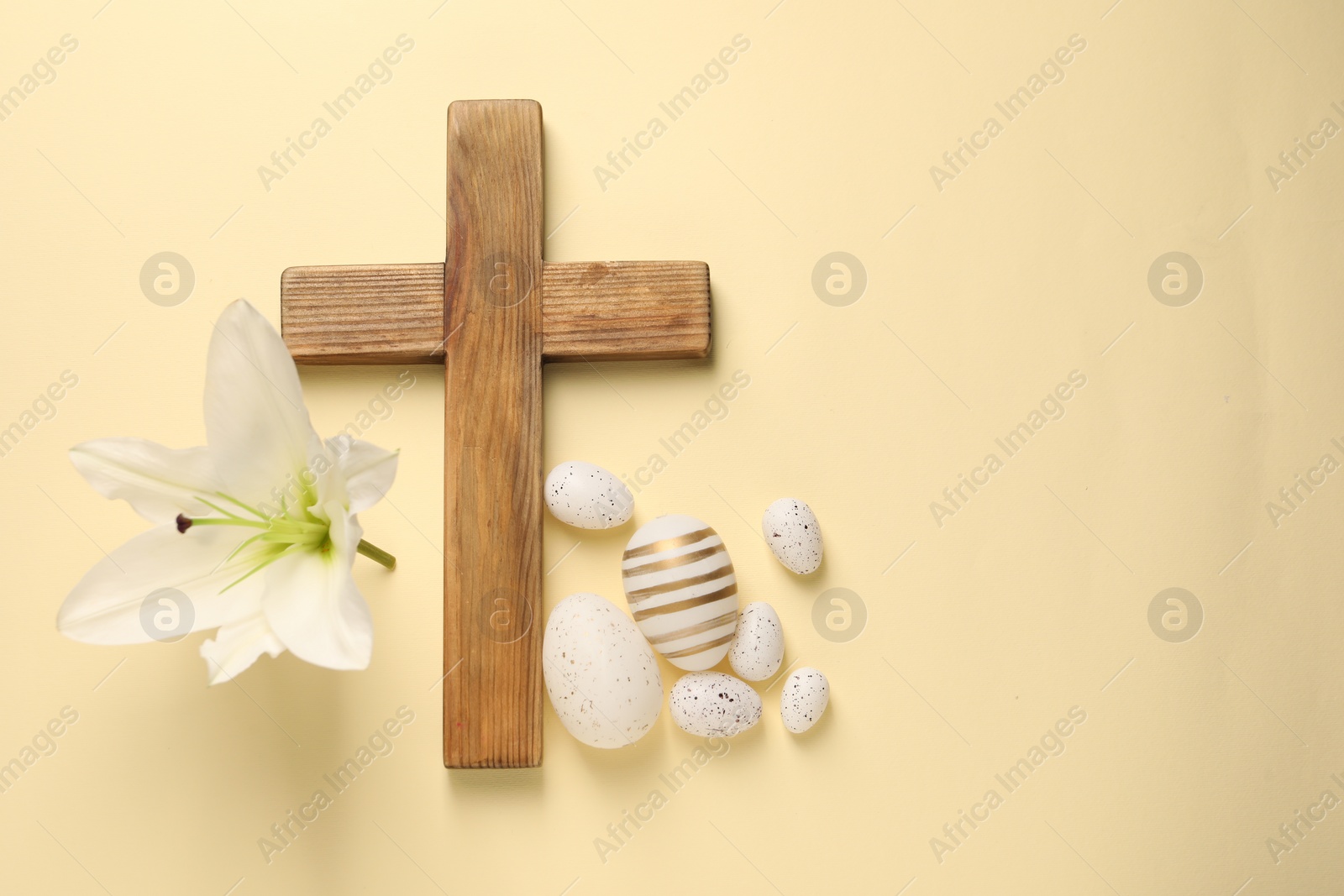 Photo of Wooden cross, painted Easter eggs and lily flower on pale yellow background, flat lay. Space for text