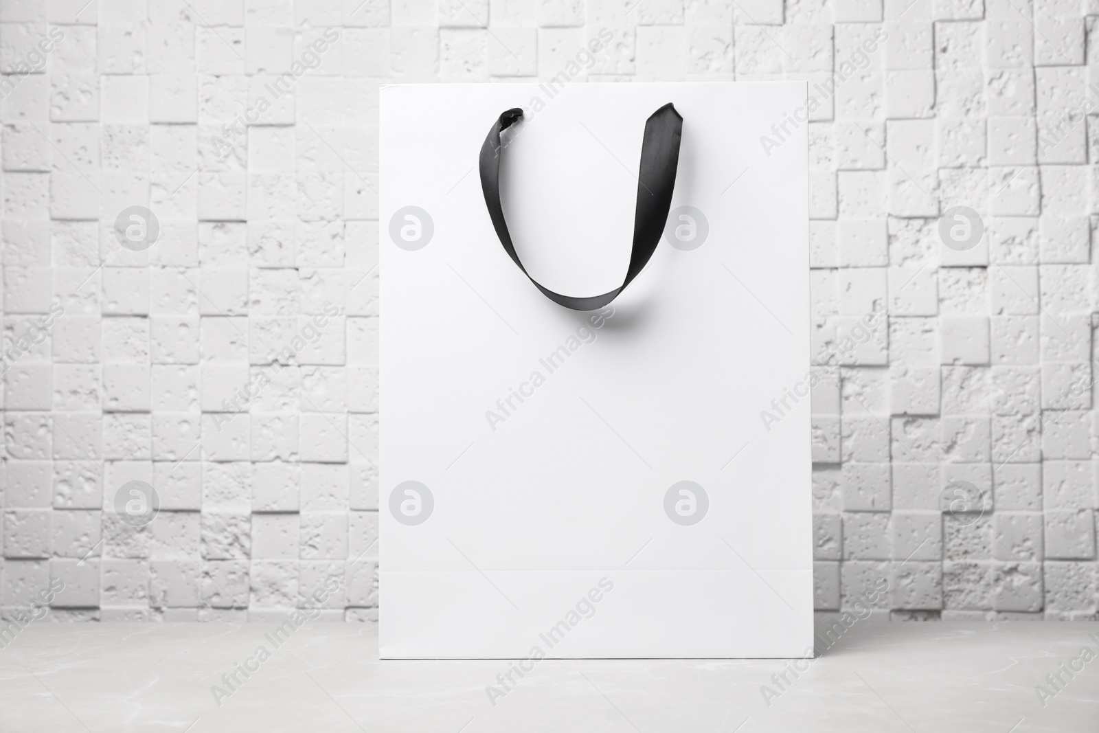 Photo of Paper shopping bag with ribbon handles on table near white wall. Mockup for design