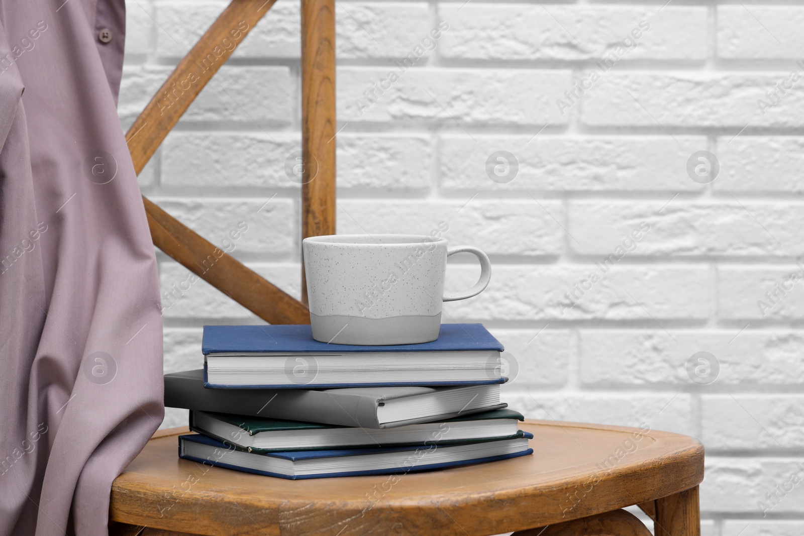 Photo of Ceramic cup and stack of books on wooden chair against white brick wall, space for text