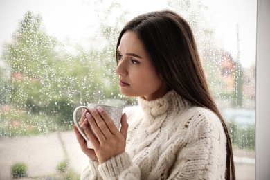 Sad beautiful woman with cup near window indoors on rainy day