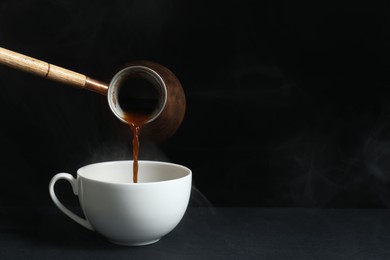 Turkish coffee. Pouring brewed beverage from cezve into cup on table against black background, space for text
