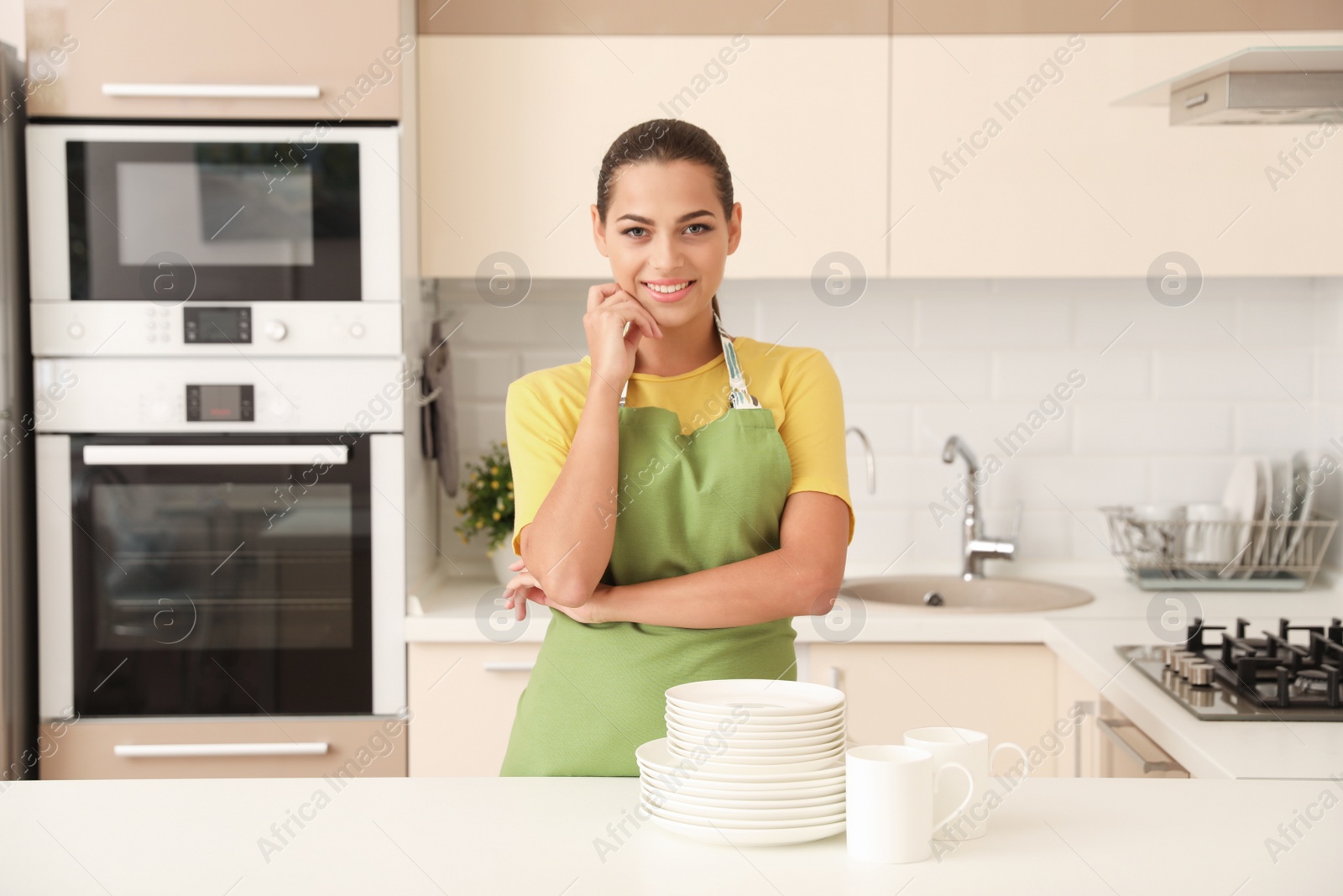 Photo of Beautiful young woman with clean dishes and cups at table in kitchen