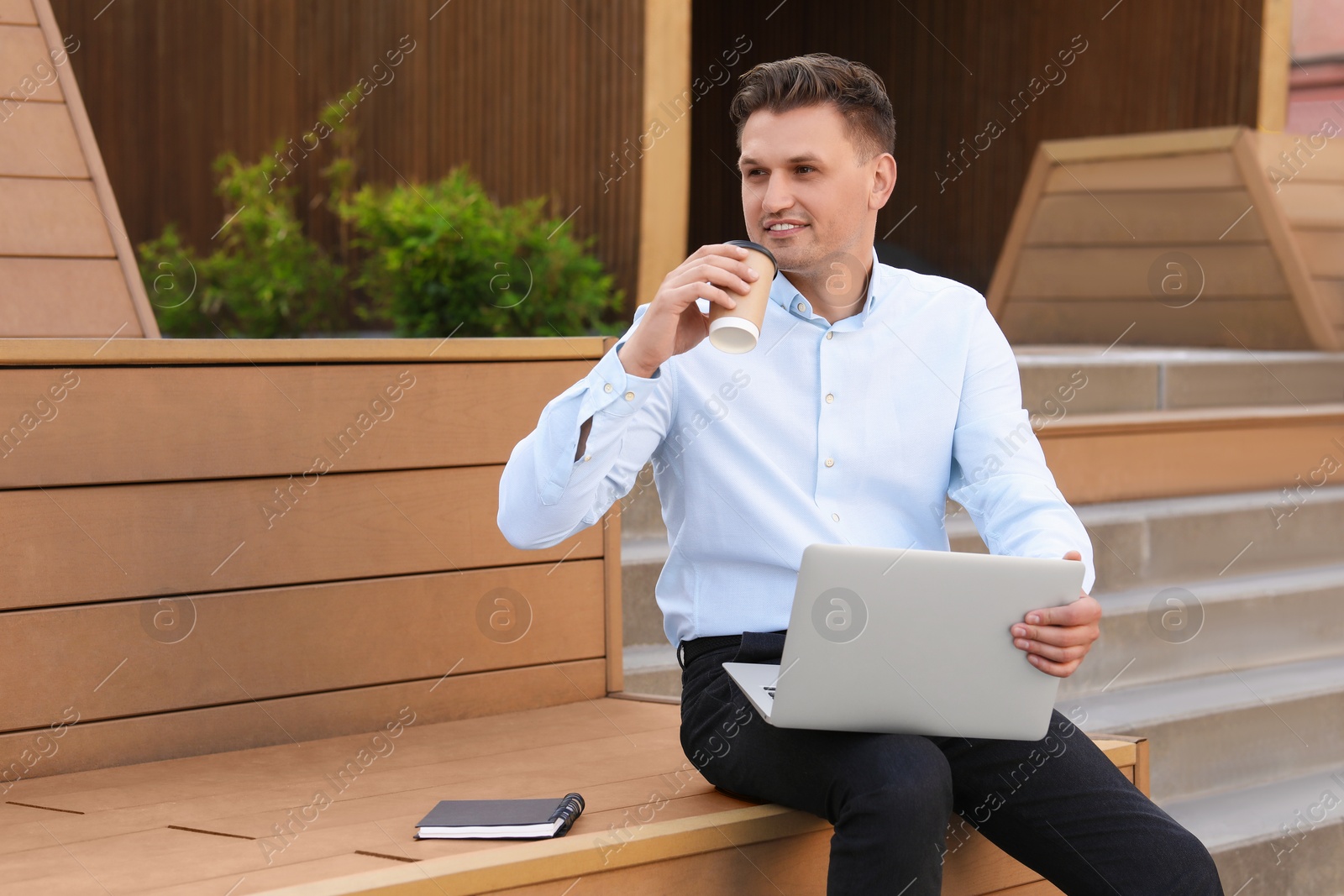 Photo of Handsome man with cup of coffee using laptop on bench outdoors. Space for text