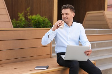 Photo of Handsome man with cup of coffee using laptop on bench outdoors. Space for text