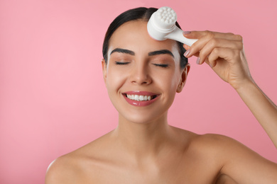 Photo of Young woman using facial cleansing brush on pink background. Washing accessory