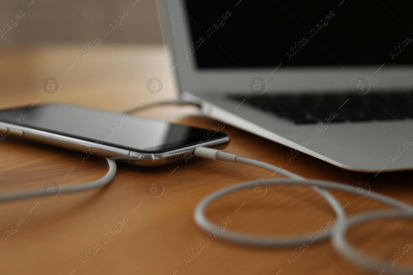 Photo of Modern smartphone charging from laptop on wooden table, closeup