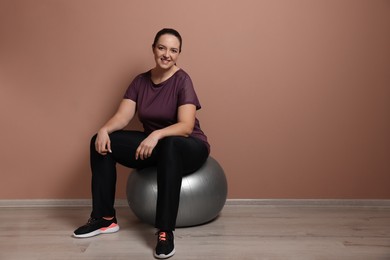 Photo of Happy overweight woman sitting on fitness ball near pale pink wall indoors. Space for text