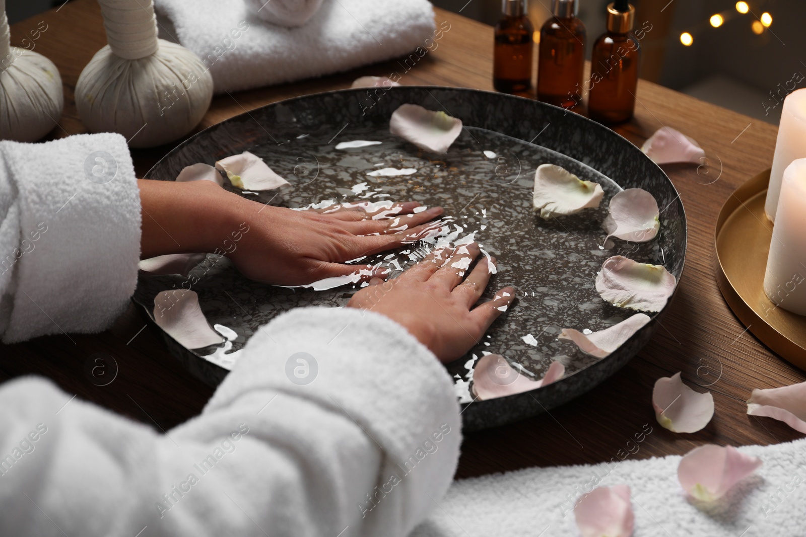 Photo of Woman soaking her hands in bowl of water and flower petals at table, closeup. Spa treatment