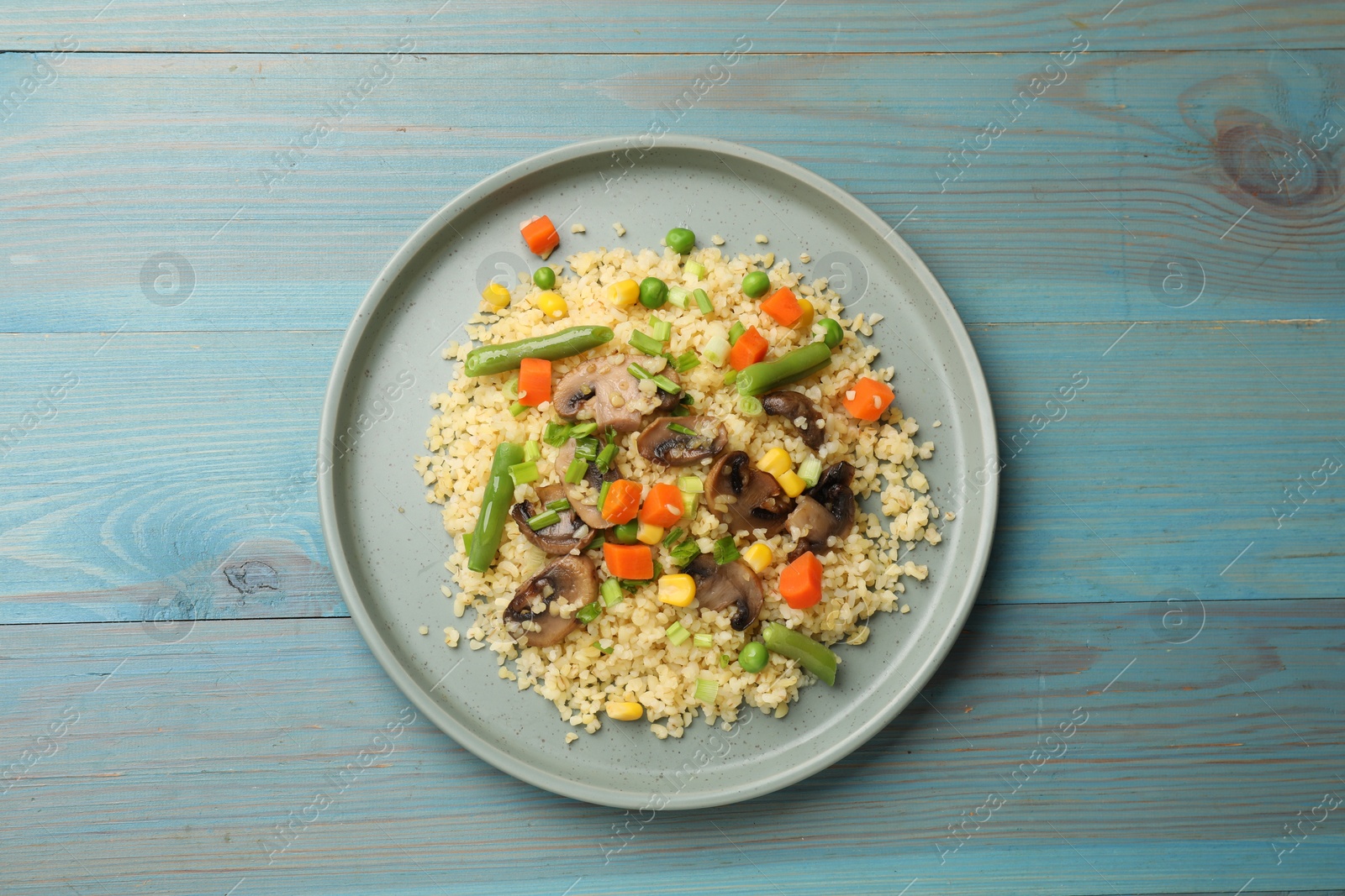 Photo of Delicious bulgur with vegetables and mushrooms on light blue wooden table, top view