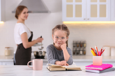 Little girl doing homework while mother cooking in kitchen