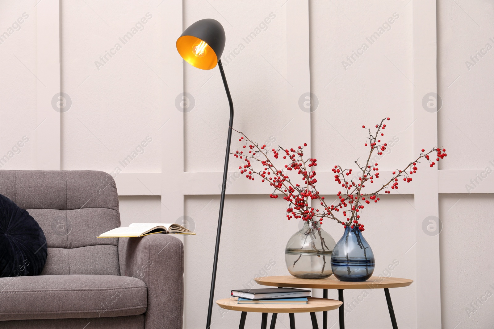 Photo of Hawthorn branches with red berries in vases, armchair and lamp indoors. Interior design