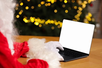 Photo of Merry Christmas. Santa Claus using laptop at table against blurred lights, closeup