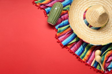 Photo of Mexican sombrero hat, toy cactus and colorful poncho on red background, flat lay. Space for text