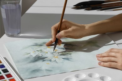 Woman painting flowers with watercolor at white table, closeup. Creative artwork