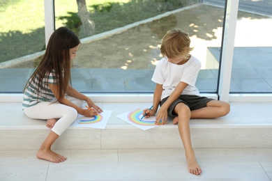 Photo of Little children drawing rainbows near window indoors. Stay at home concept