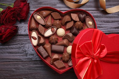 Photo of Heart shaped box with delicious chocolate candies, roses and ribbon on wooden table, flat lay