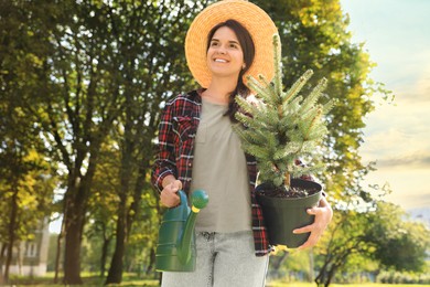 Photo of Young woman holding pot with conifer tree and watering can in park on sunny day