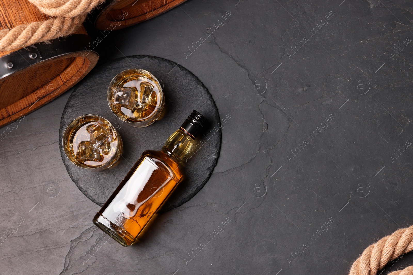 Photo of Whiskey with ice cubes in glasses, bottle, wooden barrels and rope on black table, top view. Space for text