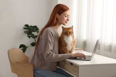 Photo of Woman working with laptop and hugging cat at desk. Home office