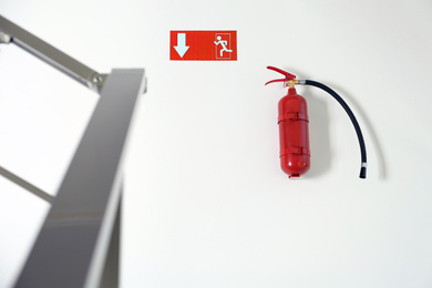 Photo of Fire extinguisher and emergency exit sign on white wall near staircase indoors