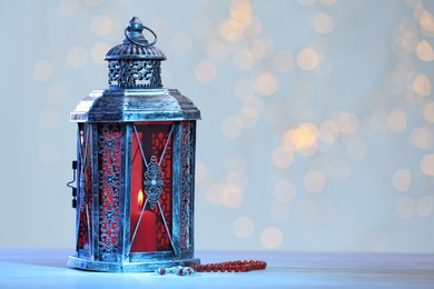 Photo of Arabic lantern and misbaha on table against blurred lights, space for text