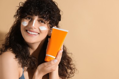 Photo of Beautiful young woman with sun protection cream on her face holding tube of sunscreen against beige background, space for text
