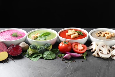 Different tasty cream soups in bowls and ingredients on dark grey table