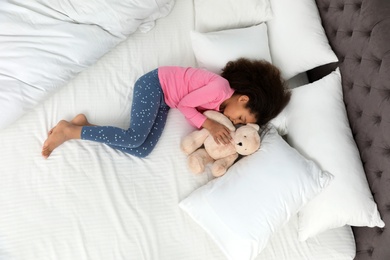 Cute little African-American girl with teddy bear sleeping in bed, top view