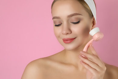 Photo of Young woman washing face with cleansing brush on pink background. Cosmetic product