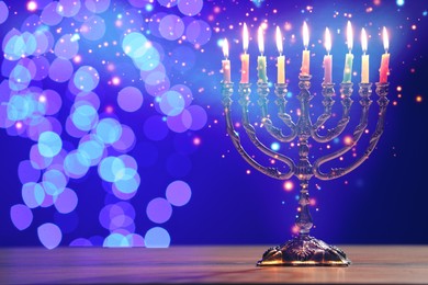 Image of Hanukkah celebration. Menorah with burning candles on wooden table against blue background with blurred lights, space for text