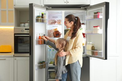 Young mother with daughter taking juice out of refrigerator in kitchen