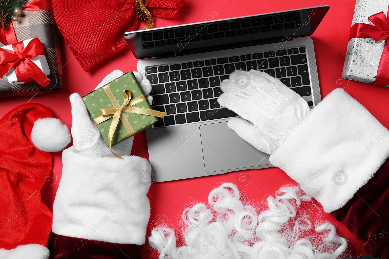 Photo of Santa Claus using laptop, closeup. Gift boxes and Christmas decor on red background, top view