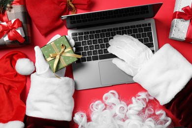 Santa Claus using laptop, closeup. Gift boxes and Christmas decor on red background, top view
