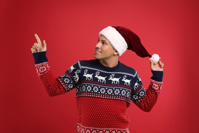Photo of Handsome man in Christmas sweater and Santa hat pointing on red background